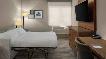 Ein Hotelzimmer mit Bett, Schreibtisch, Stuhl, Fernseher, Lampe, Klimaanlage und gerahmten Bildern an der Wand.