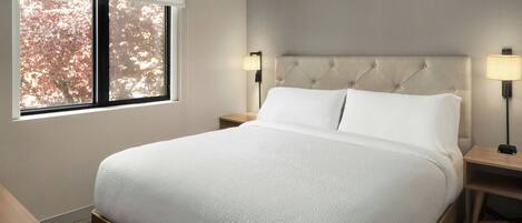 A hotel room with a large bed, two bedside tables, a window with a view of trees, and a wall-mounted lamp.
