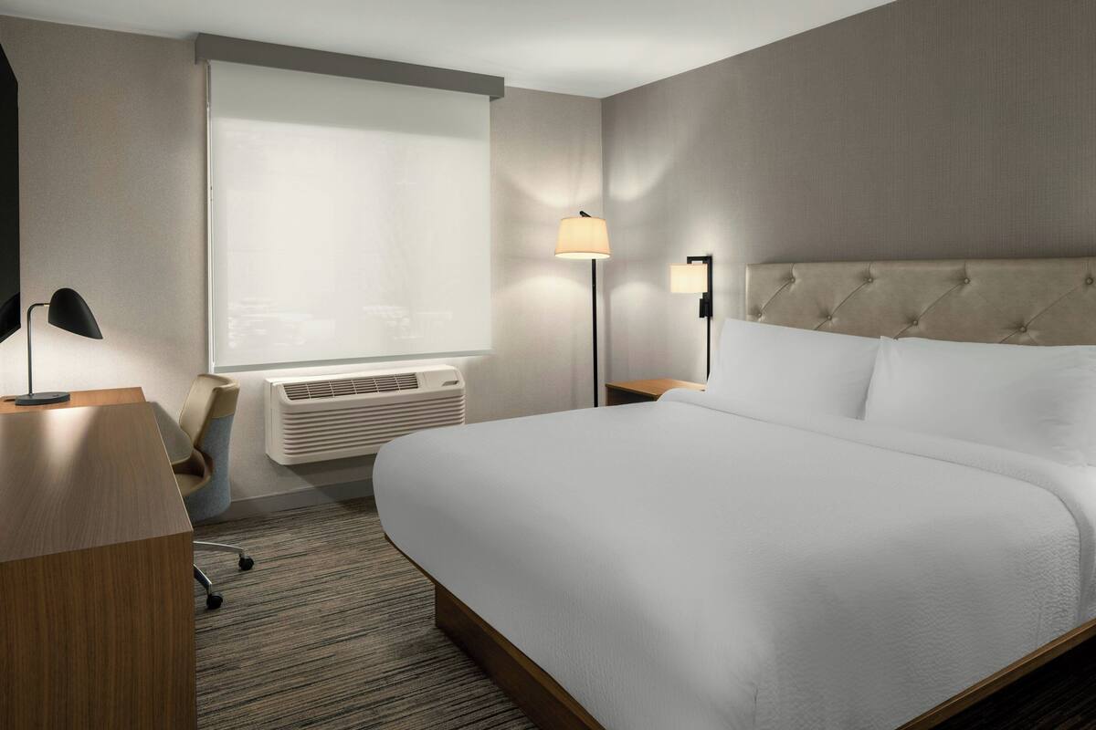 Habitación de hotel con una cama grande, un escritorio, una silla, una lámpara de pie y una ventana con persianas.