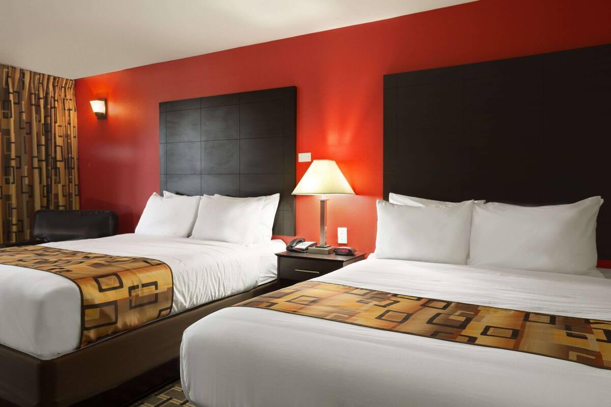 A hotel room with two beds, a red accent wall, a patterned throw, and a nightstand with a lamp.