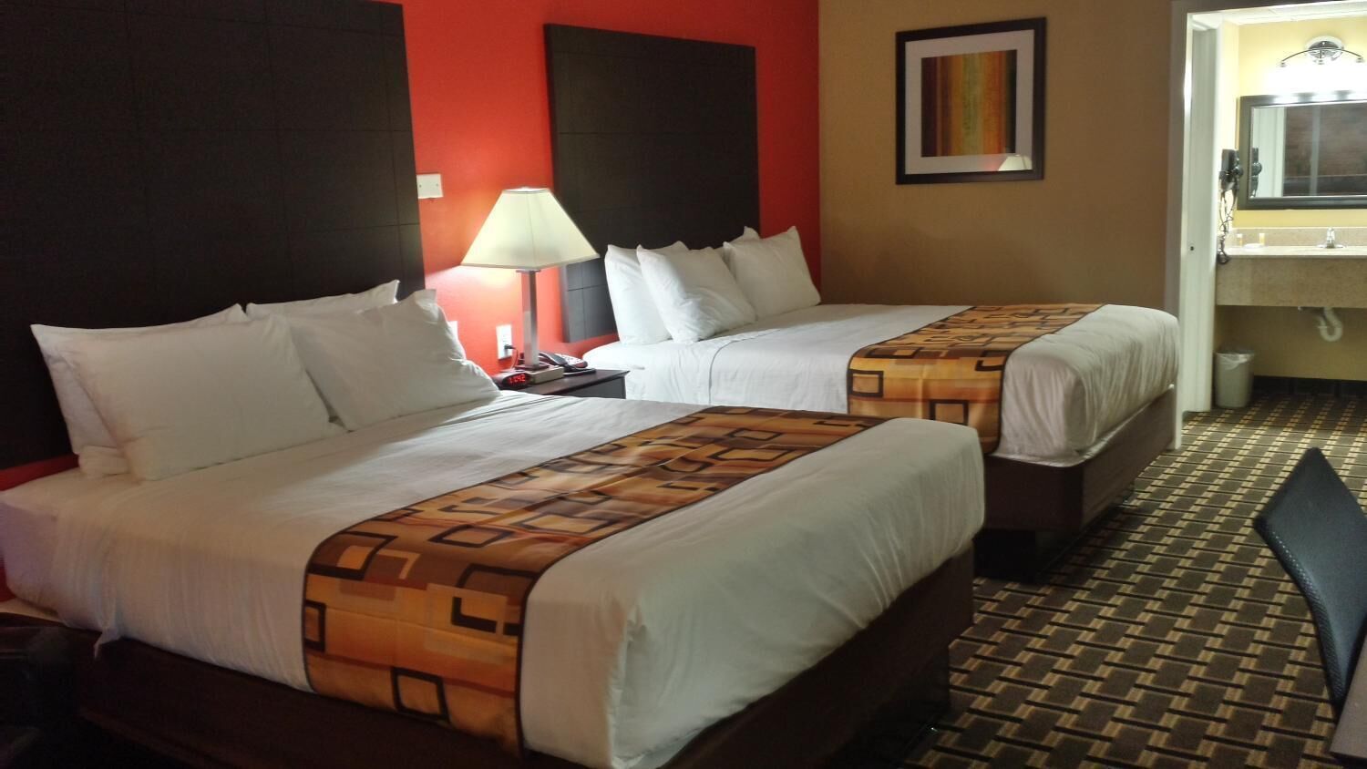 A hotel room with two beds, a nightstand with a lamp, a bathroom with a mirror, and a patterned floor.