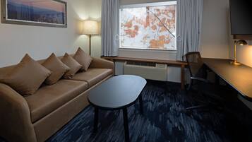 Quarto de hotel com sofá, mesa pequena, escrivaninha, cadeira, televisão, janela com cortinas e abajur.