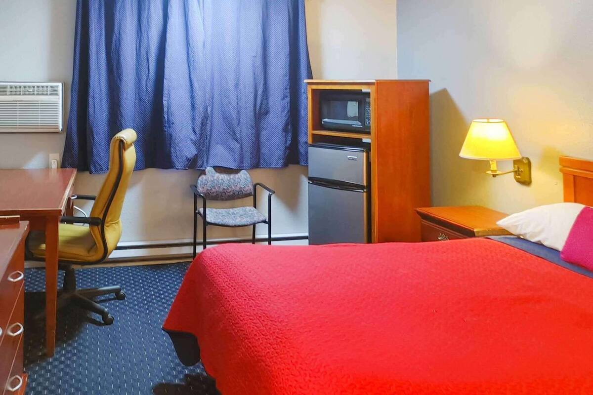 Ein Hotelzimmer mit Bett, Schreibtisch, Stuhl, Mikrowelle, Kühlschrank, Lampe und einem Fenster mit Vorhängen.