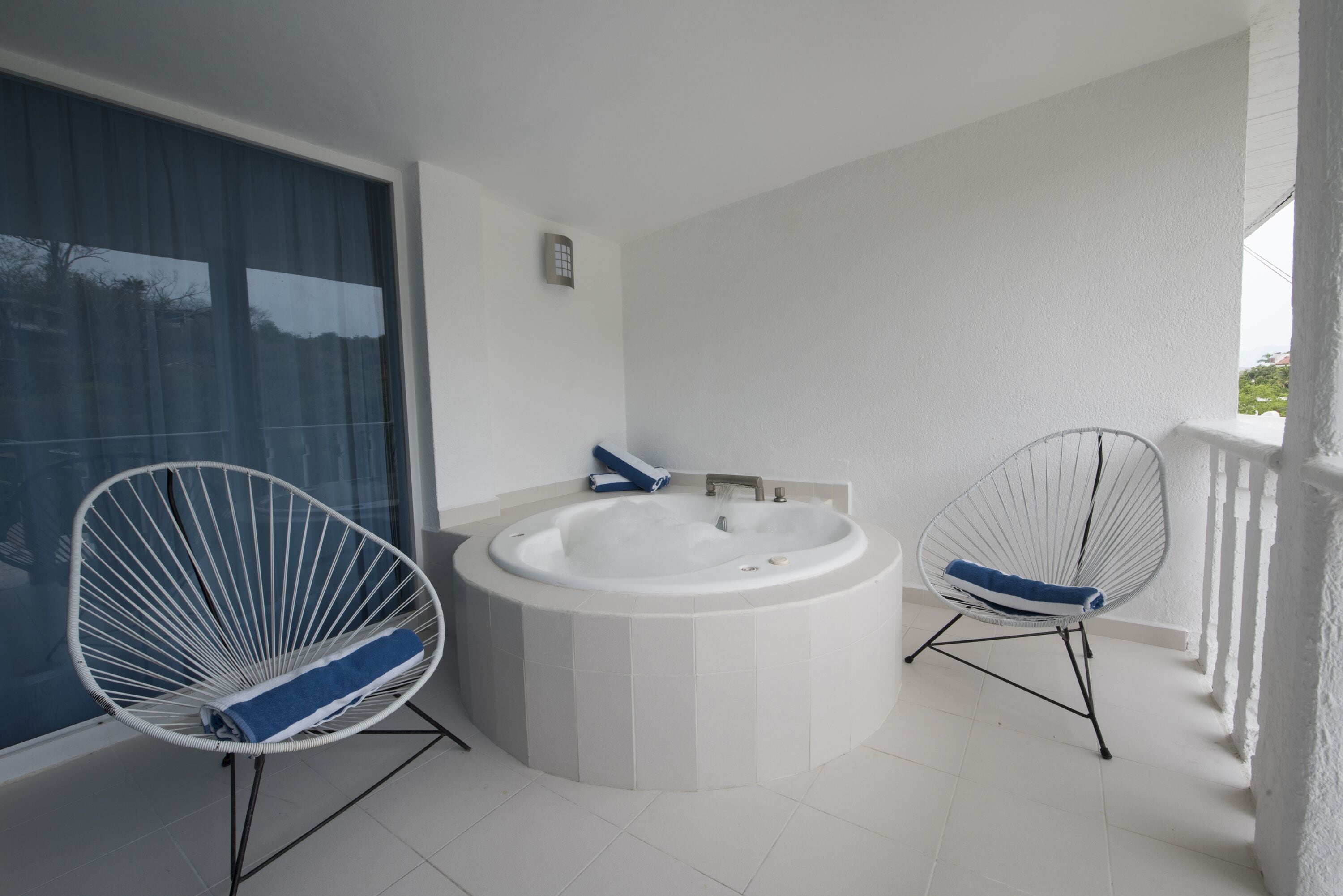 A modern bathroom with a freestanding bathtub, two wire-framed chairs, and a large window with curtains.
