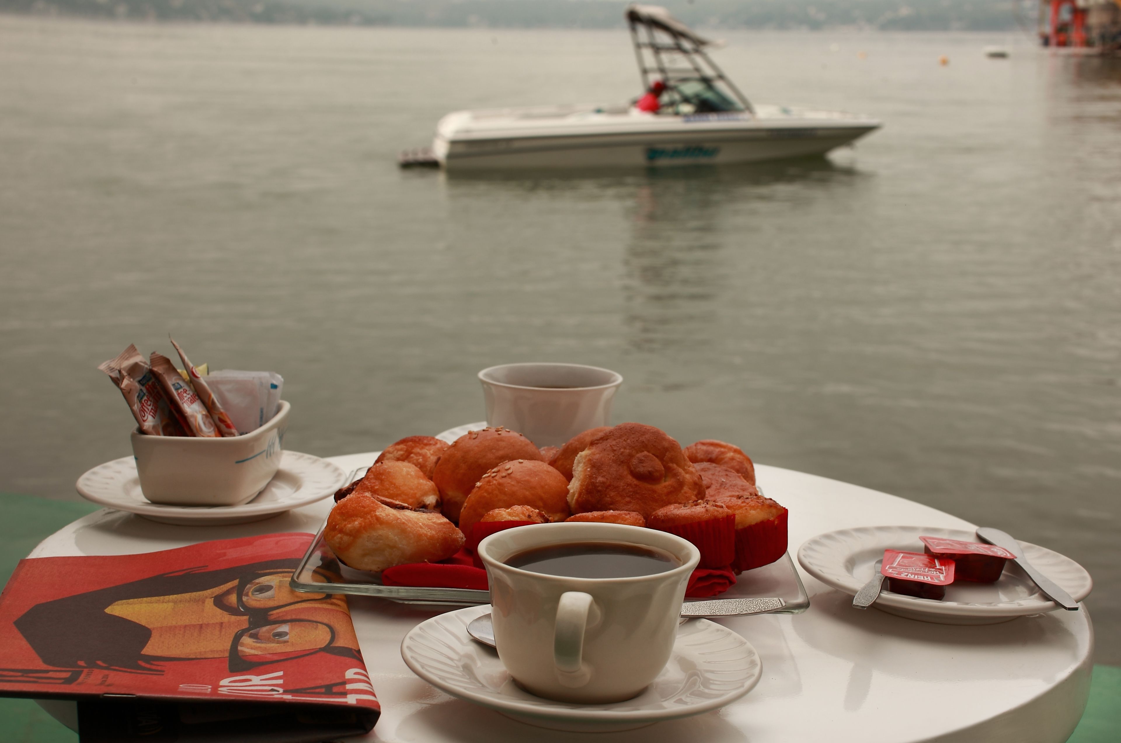 Une table avec des pâtisseries, une tasse de café et une assiette de nourriture, le tout placé près d’un lac, avec un bateau en arrière-plan.
