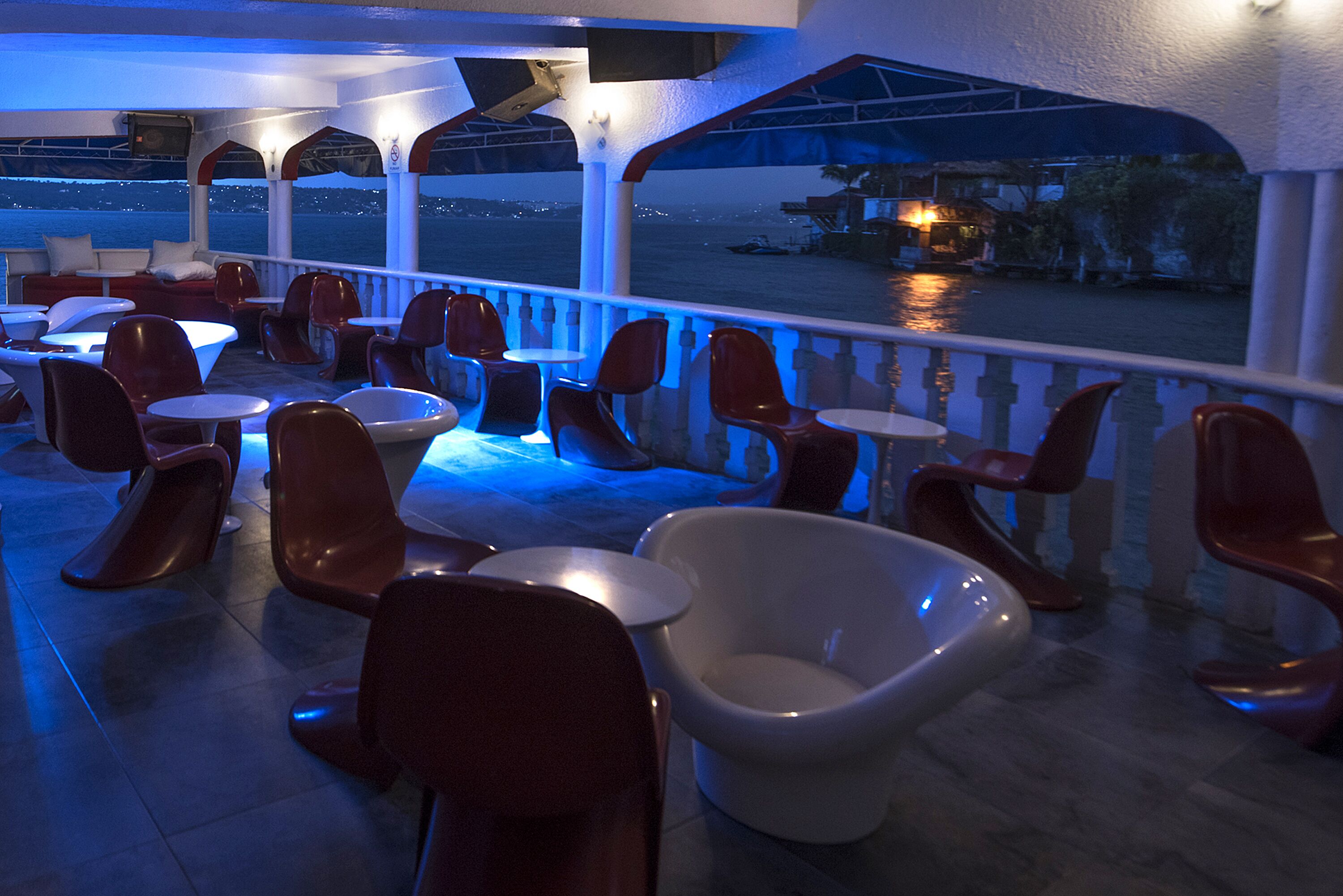 Un restaurante con vistas al agua, con sillas rojas, mesas blancas e iluminación azul en el techo.