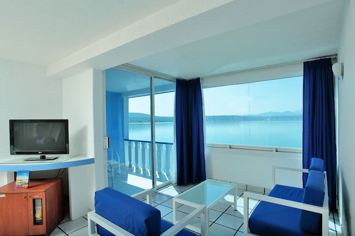A room with a TV, a sofa, a glass table, and a balcony with a view of the sea.