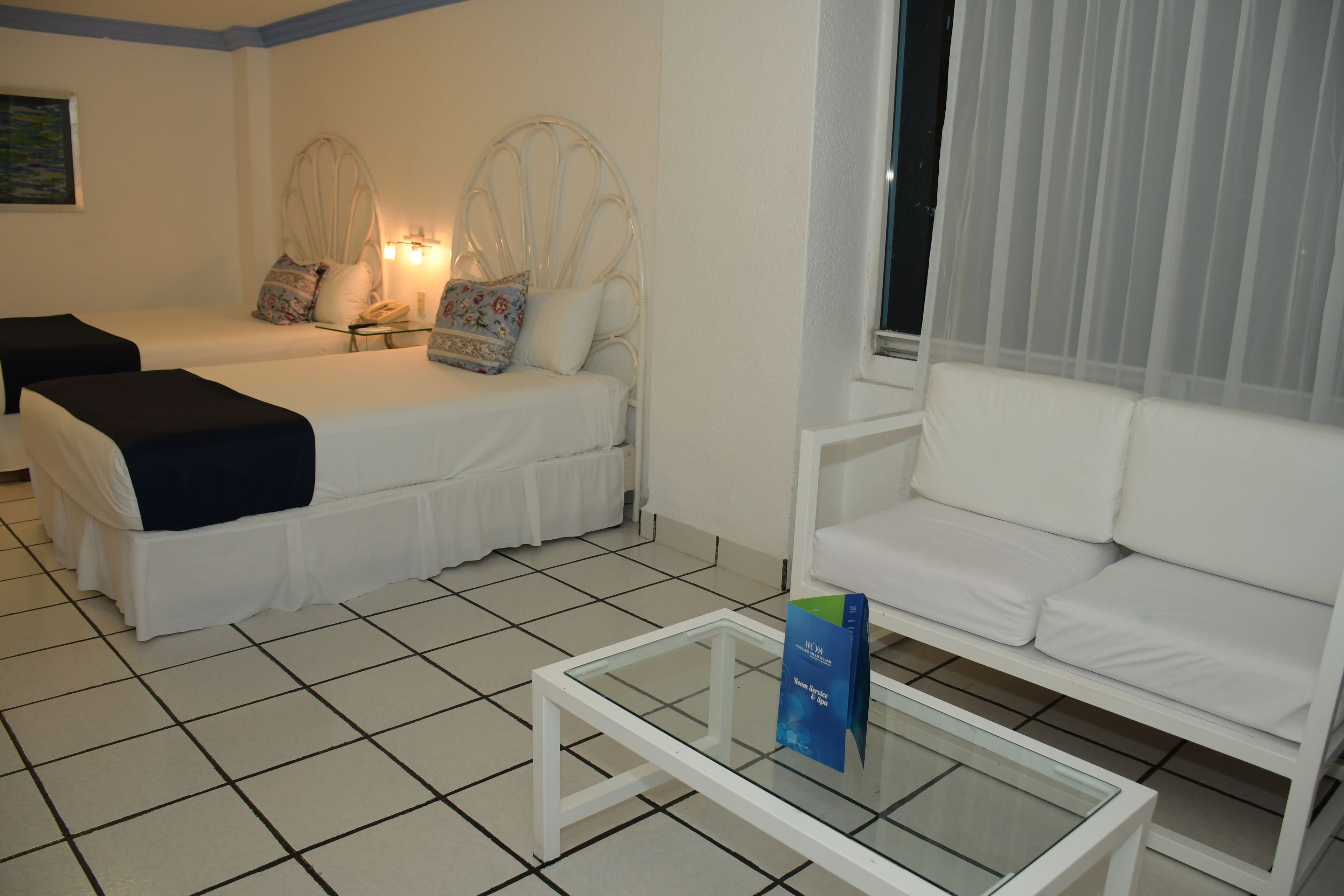 Habitación de hotel con dos camas, un sofá, una mesita de centro de cristal y una ventana con cortinas de encaje.
