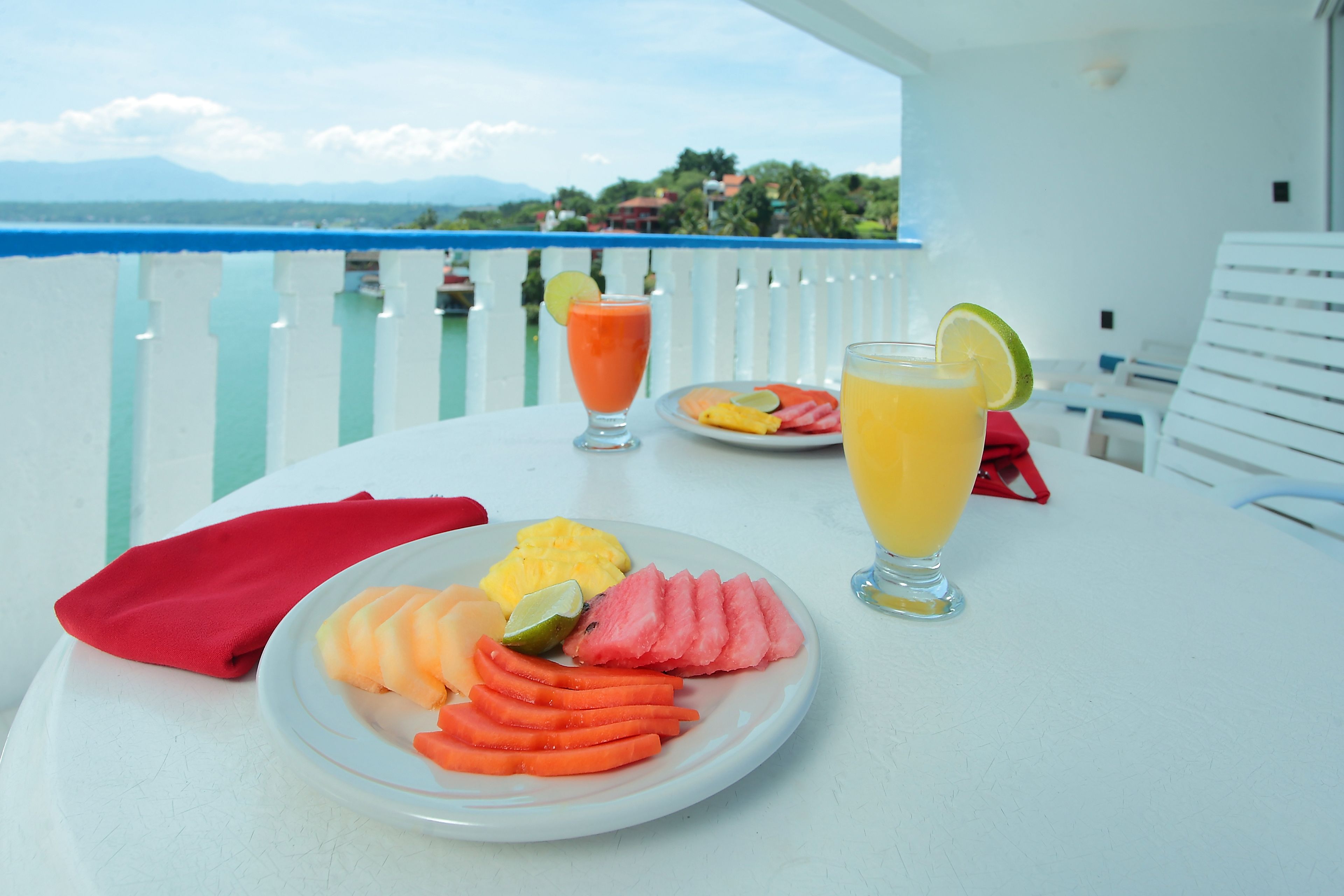 A table with a plate of fruit and two drinks, a red napkin, and a white chair on a balcony with a view of the sea and mountains.