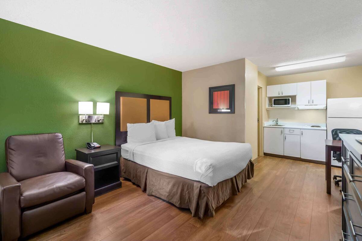 A hotel room with a bed, a desk, a chair, a nightstand, a lamp, a microwave, and cabinets.