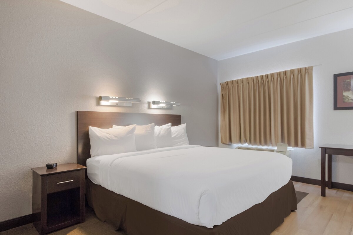 A hotel room with a large bed, bedside tables, a nightstand, a window with curtains, and a wall-mounted light fixture.