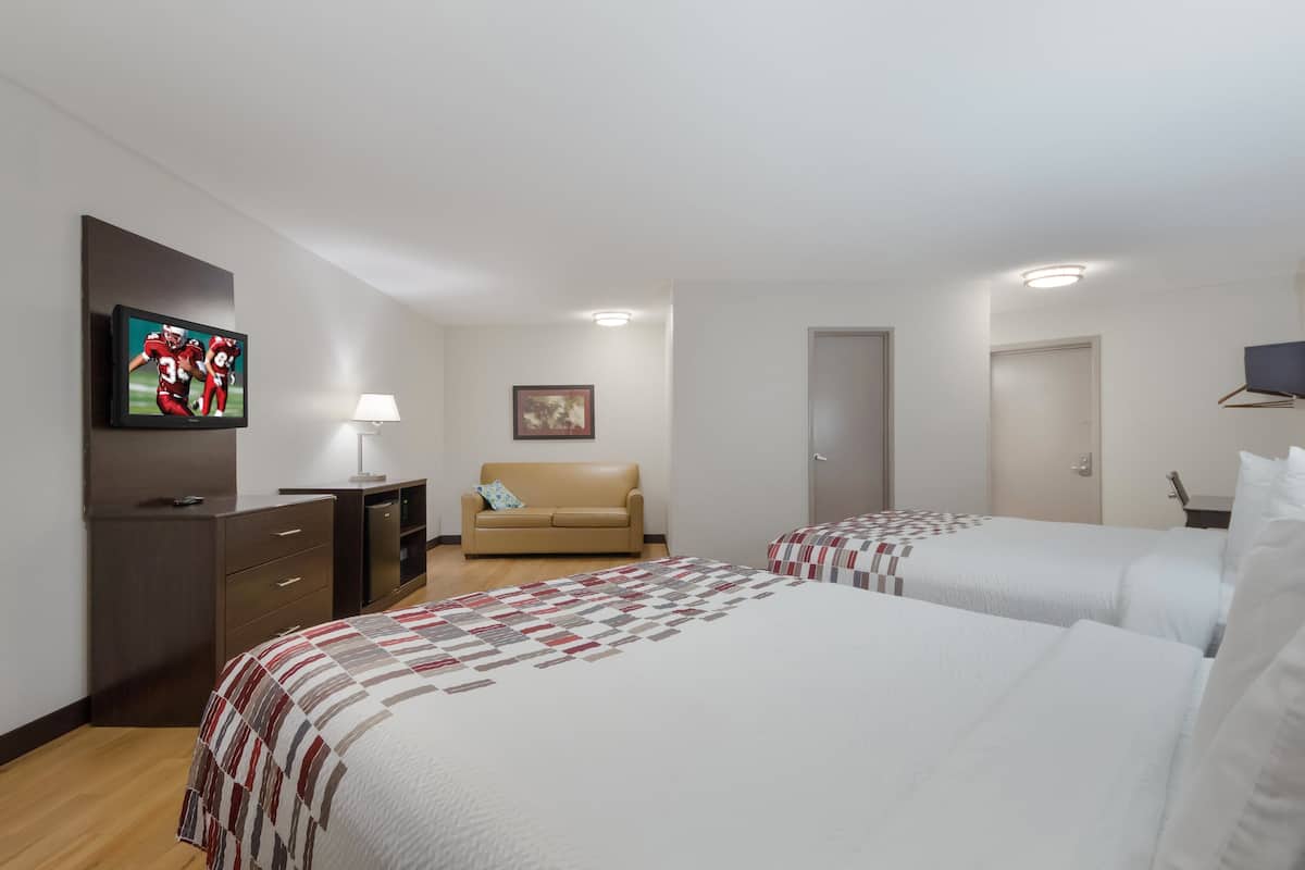 Une chambre d’hôtel avec deux lits, un téléviseur fixé au mur, un bureau avec une lampe, un canapé et un tableau au mur.