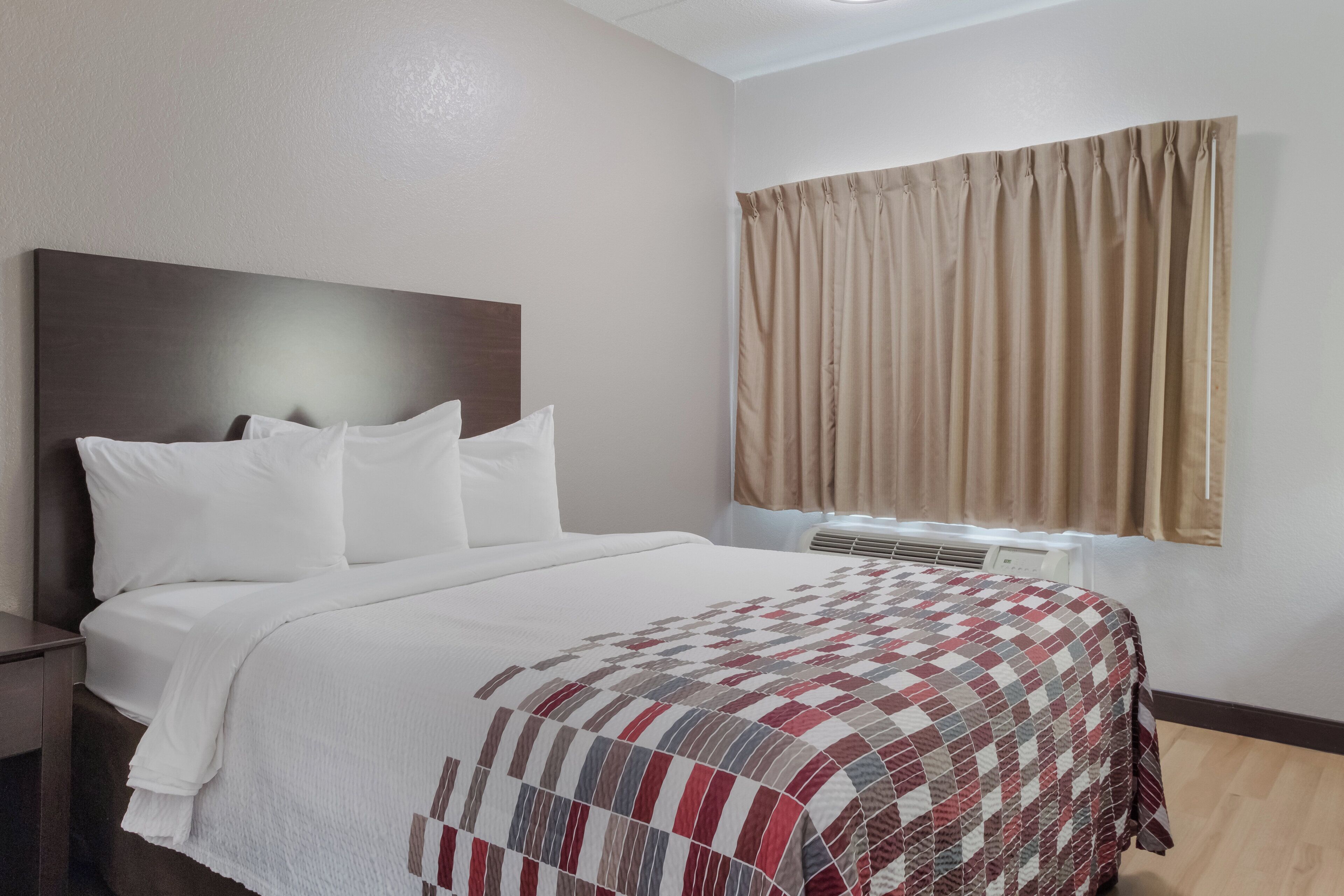 A hotel room with a bed, a nightstand, a window with curtains, and a wall-mounted air conditioning unit.