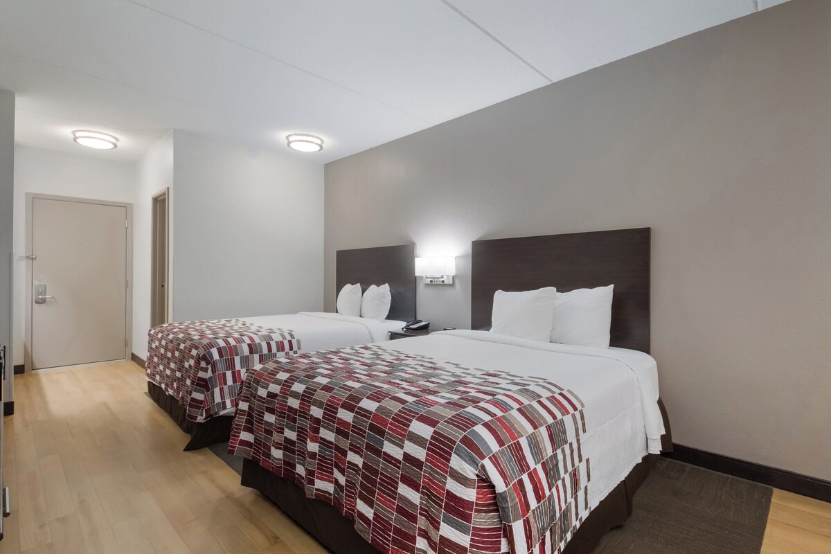 A hotel room with two beds, each with a patterned bedspread, a bedside table with a lamp, and a door leading to another room.