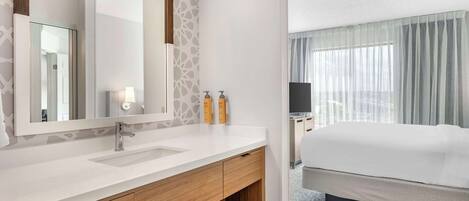 A hotel room with a white bathroom vanity, a large mirror, a wooden cabinet, and a bed with a white comforter.