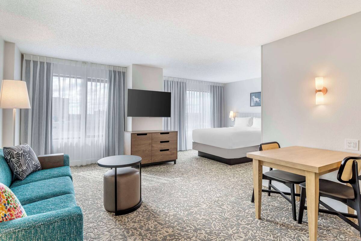 A hotel room with a bed, a desk, a chair, a TV, a lamp, and a sofa.