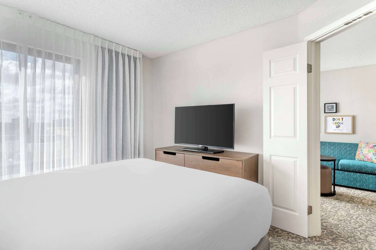 A hotel room with a bed, a TV on a wooden dresser, a door leading to another room, and a framed motivational quote on the wall.