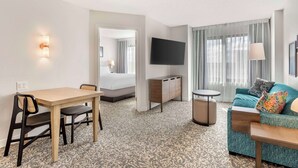 Habitación de hotel con una mesa de comedor de madera, un sofá con cojines coloridos, un televisor de pantalla plana en la pared y una alfombra estampada.
