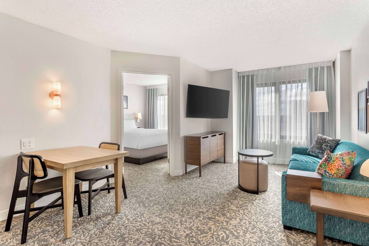 A hotel room with a wooden dining table, a sofa with colorful pillows, a flat-screen TV mounted on the wall, and a patterned carpet.