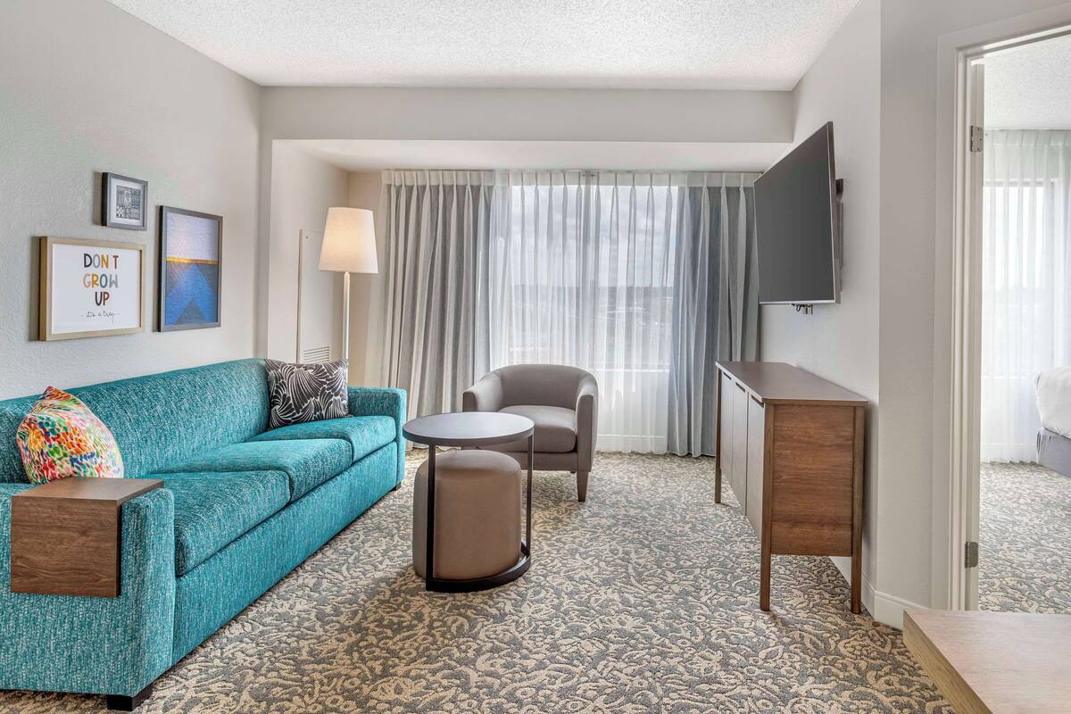 A hotel room with a teal sofa, a round ottoman, a grey armchair, a small round table, a TV mounted on the wall, and a patterned carpet.