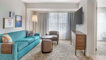 A hotel room with a teal sofa, a round ottoman, a grey armchair, a small round table, a TV mounted on the wall, and a patterned carpet.