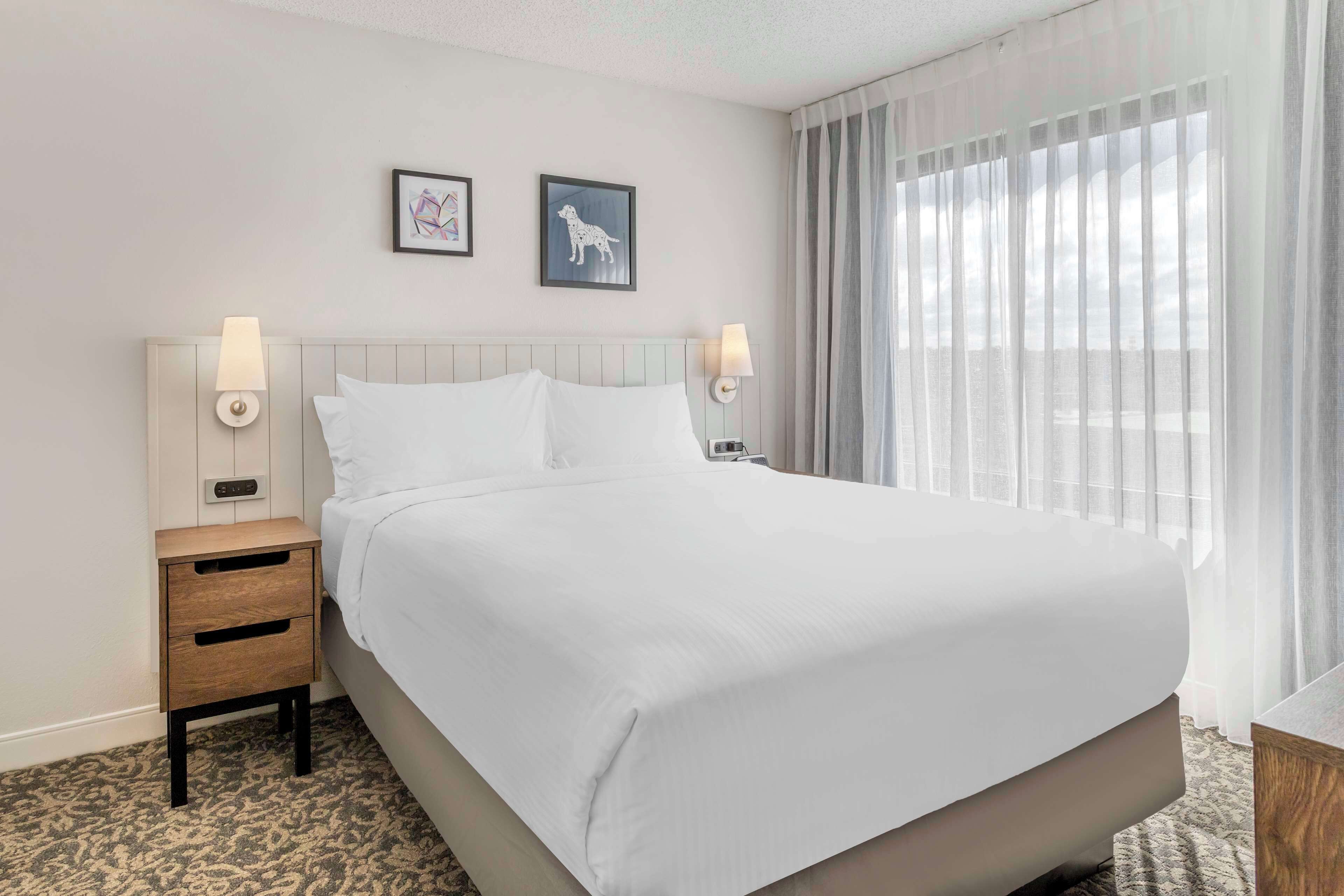 A neatly made bed with white linens, a wooden nightstand with drawers, two wall-mounted lamps, and two framed pictures on the wall.