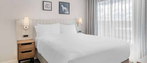 A neatly made bed with white linens, a wooden nightstand with drawers, two wall-mounted lamps, and two framed pictures on the wall.
