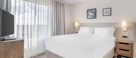 A hotel room with a large bed, a TV on a wooden cabinet, two wall-mounted lamps, and a window with sheer curtains.