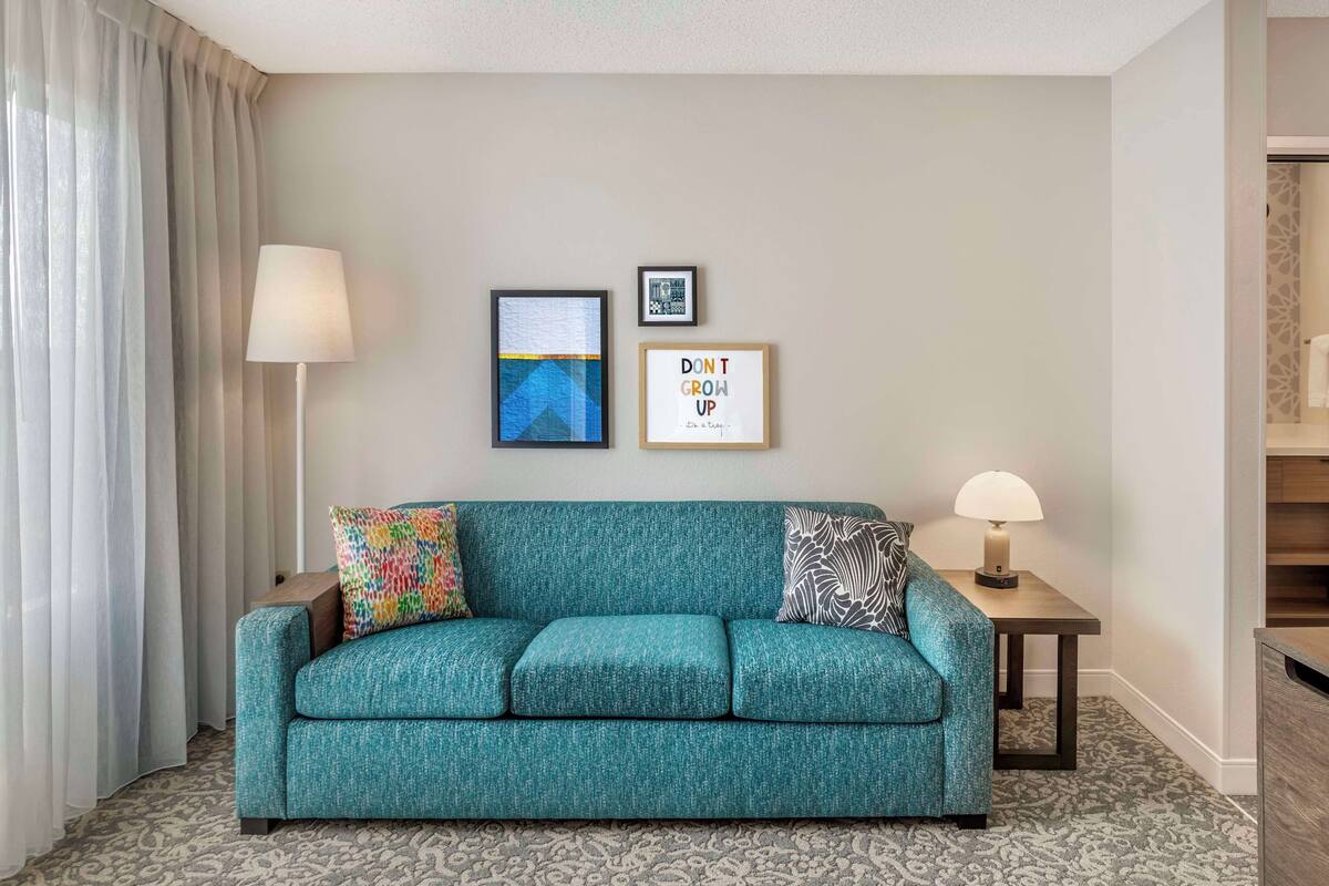 A teal sofa with patterned cushions, a small table with a lamp, and two framed pictures on the wall.