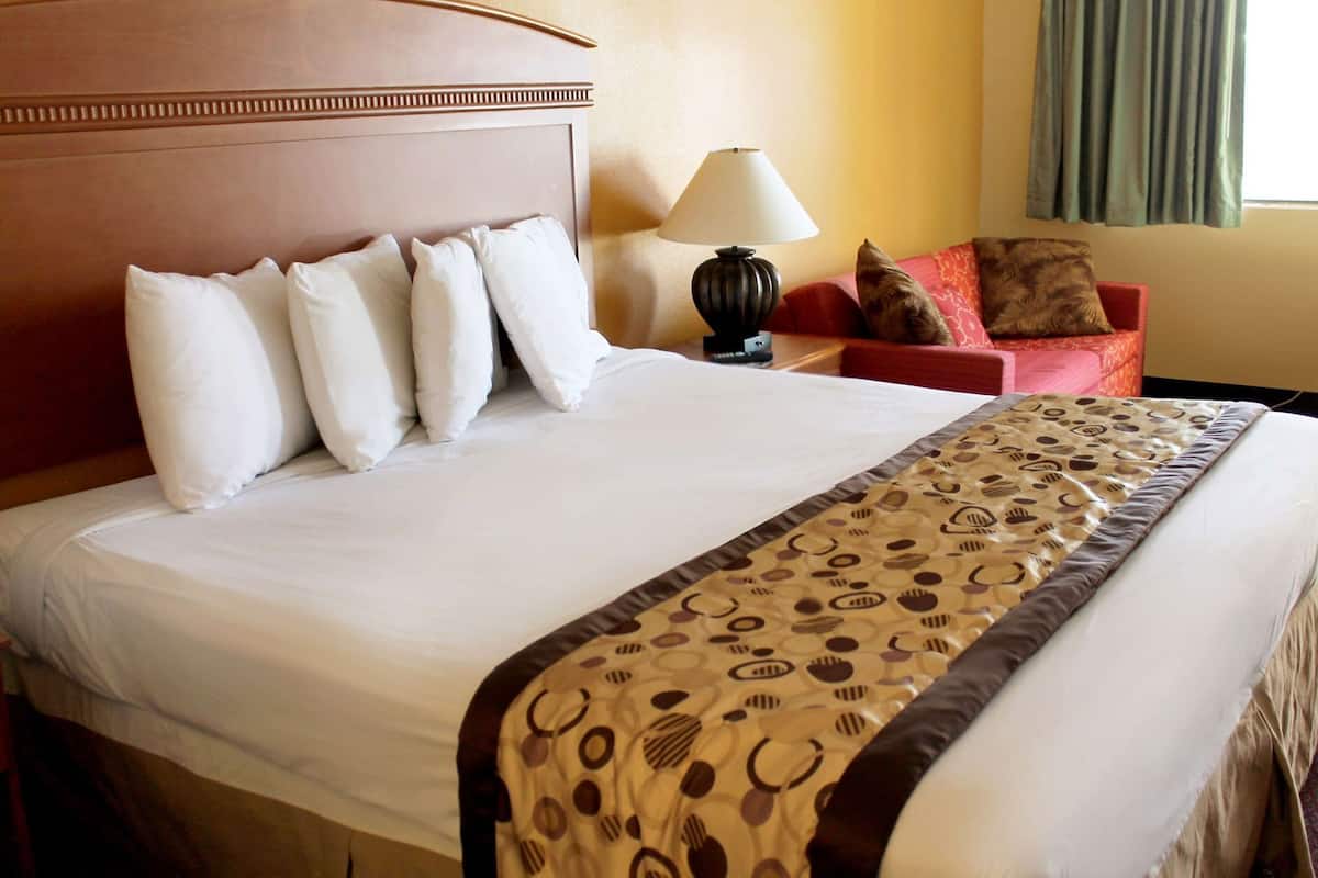 Une chambre d’hôtel avec un grand lit, une table de chevet avec une lampe, un fauteuil rouge et une fenêtre avec des rideaux.
