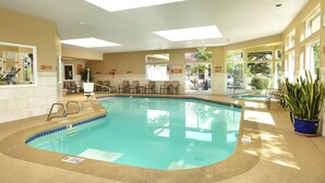 Una piscina interior con borde de azulejos, tragaluces y una zona de estar con sillas y mesas.