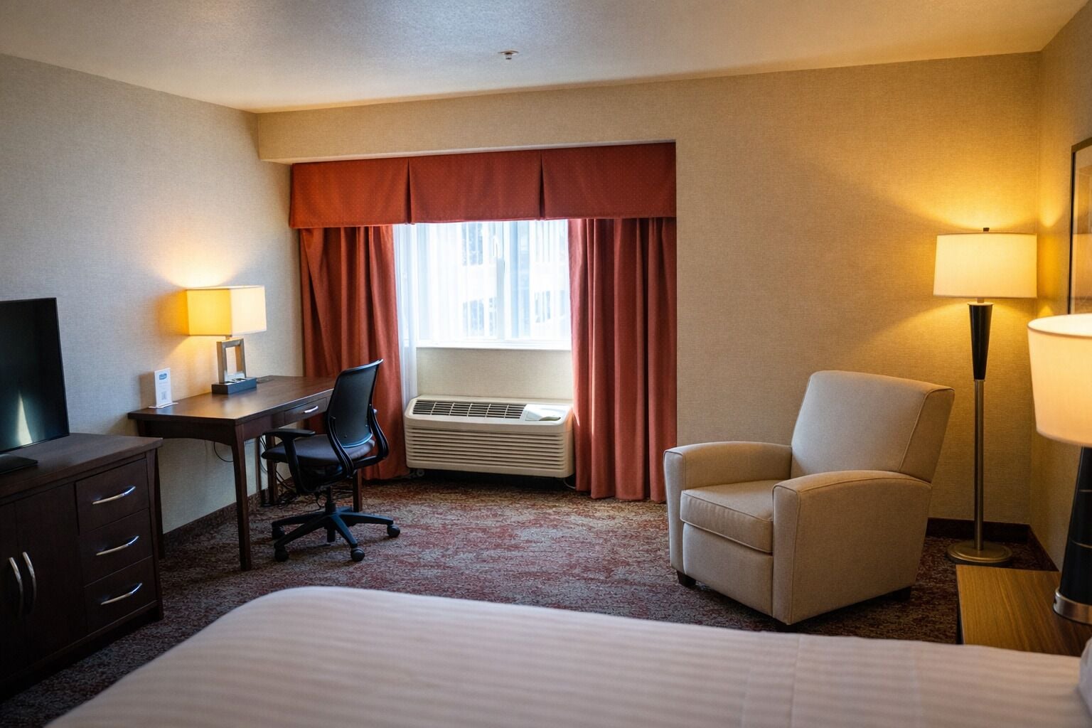 A hotel room with a bed, a desk, a chair, a TV, an air conditioner, and a lamp.
