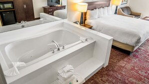 Habitación de hotel con una bañera blanca grande, una cama con ropa blanca, un armario de madera, un televisor sobre un mueble de madera y dos lámparas de noche.