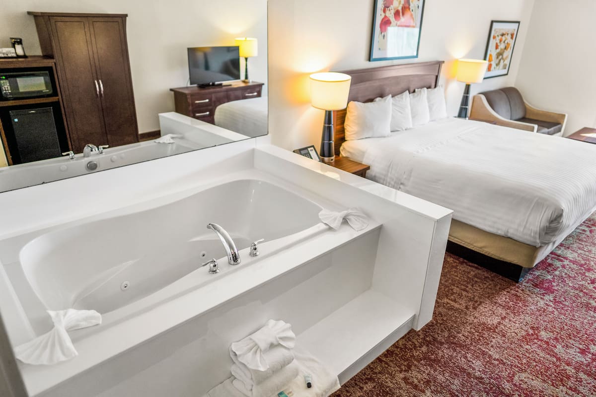 A hotel room with a large white bathtub, a bed with white linens, a wooden wardrobe, a television on a wooden stand, and two bedside lamps.