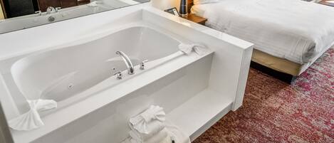Habitación de hotel con una bañera blanca grande, una cama con ropa blanca, un armario de madera, un televisor sobre un mueble de madera y dos lámparas de noche.