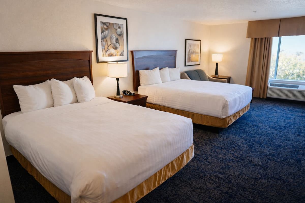 A hotel room with two beds, a nightstand with a lamp, a telephone, and a window with curtains.