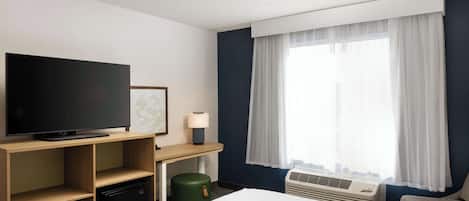 Una habitación de hotel con cama, televisión de pantalla plana, escritorio con lámpara, cesto de basura verde y ventana con cortinas.