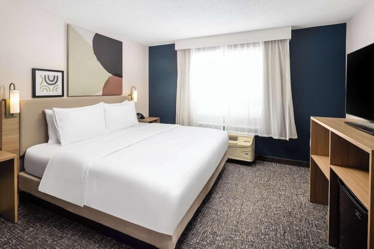 A hotel room with a bed, bedside tables, a TV, a window with curtains, and artwork on the wall.
