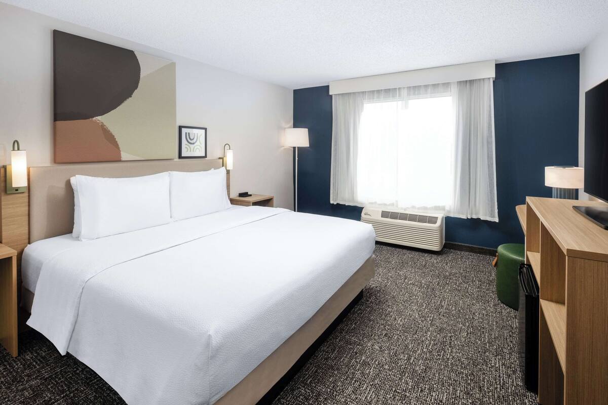 A hotel room with a large bed, two bedside tables, a desk, a chair, a flat-screen TV, a window with curtains, an air conditioning unit, and a wall-mounted artwork.