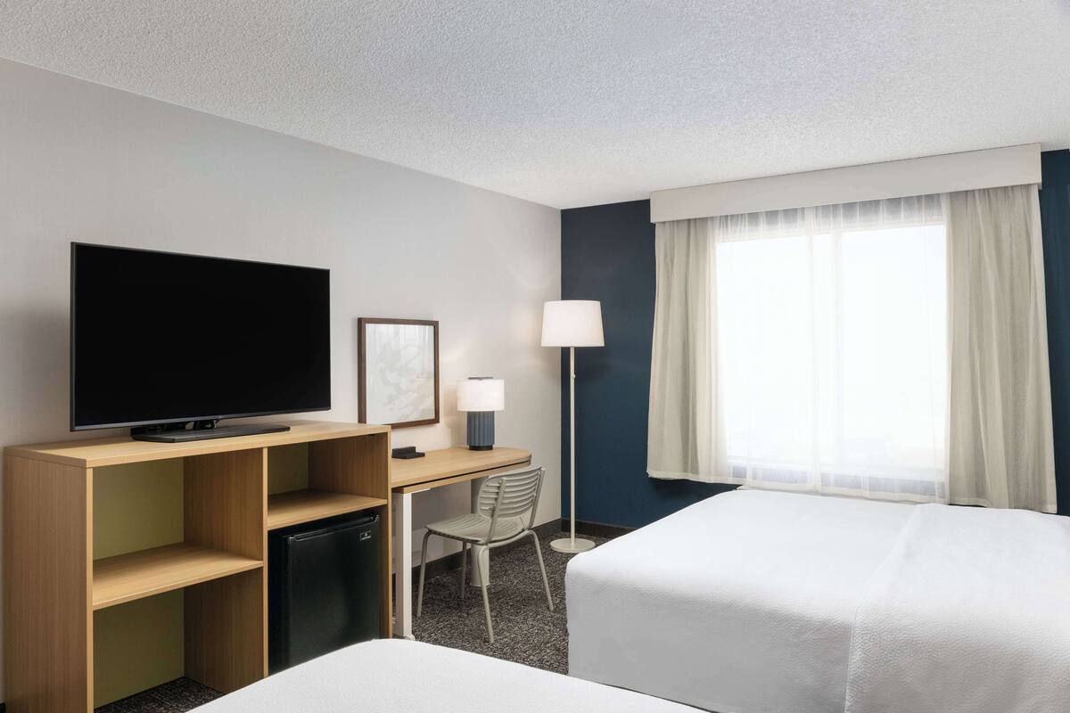 A hotel room with a flat-screen TV, a desk with a lamp, a chair, and two beds.