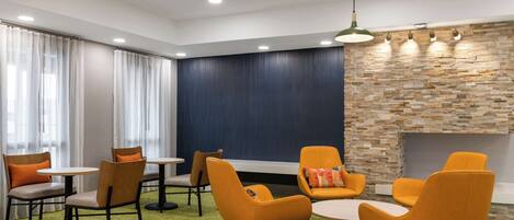 Un área de descanso moderna con sillas color naranja, mesas redondas, una lámpara colgante verde y una pared de piedra.