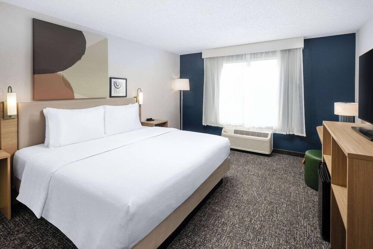 A hotel room with a large bed, a desk, a chair, a flat-screen TV, a window with curtains, an air conditioning unit, and a wall-mounted artwork.
