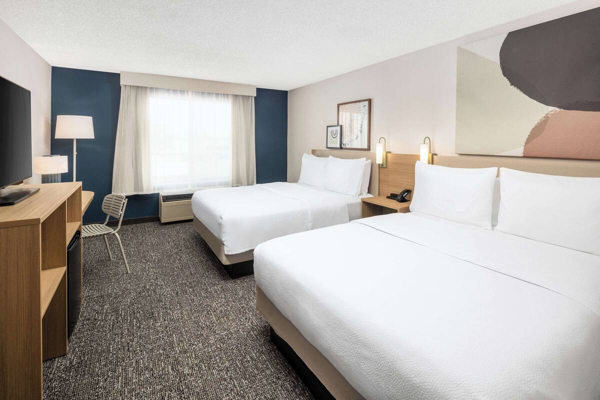 A hotel room with two beds, a desk, a chair, a TV, and a large window with curtains.