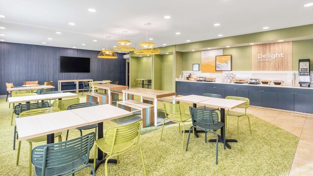 A modern dining area with multiple tables and chairs, a kitchen counter, and a flat-screen TV mounted on the wall. - Spark by Hilton Salt Lake City Airport (Salt Lake City)