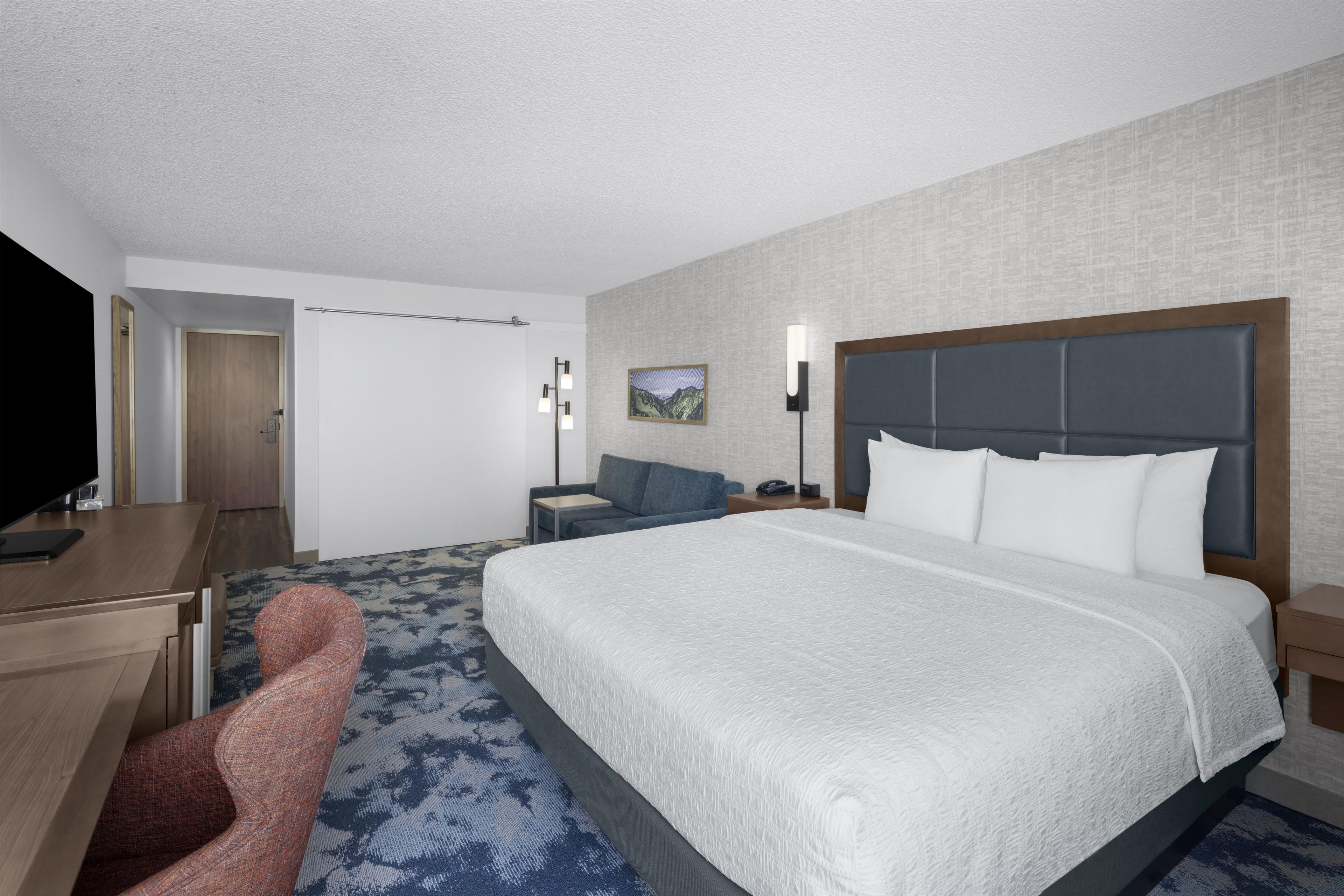 A hotel room with a large bed, a desk, a chair, a nightstand, and a wall-mounted TV.