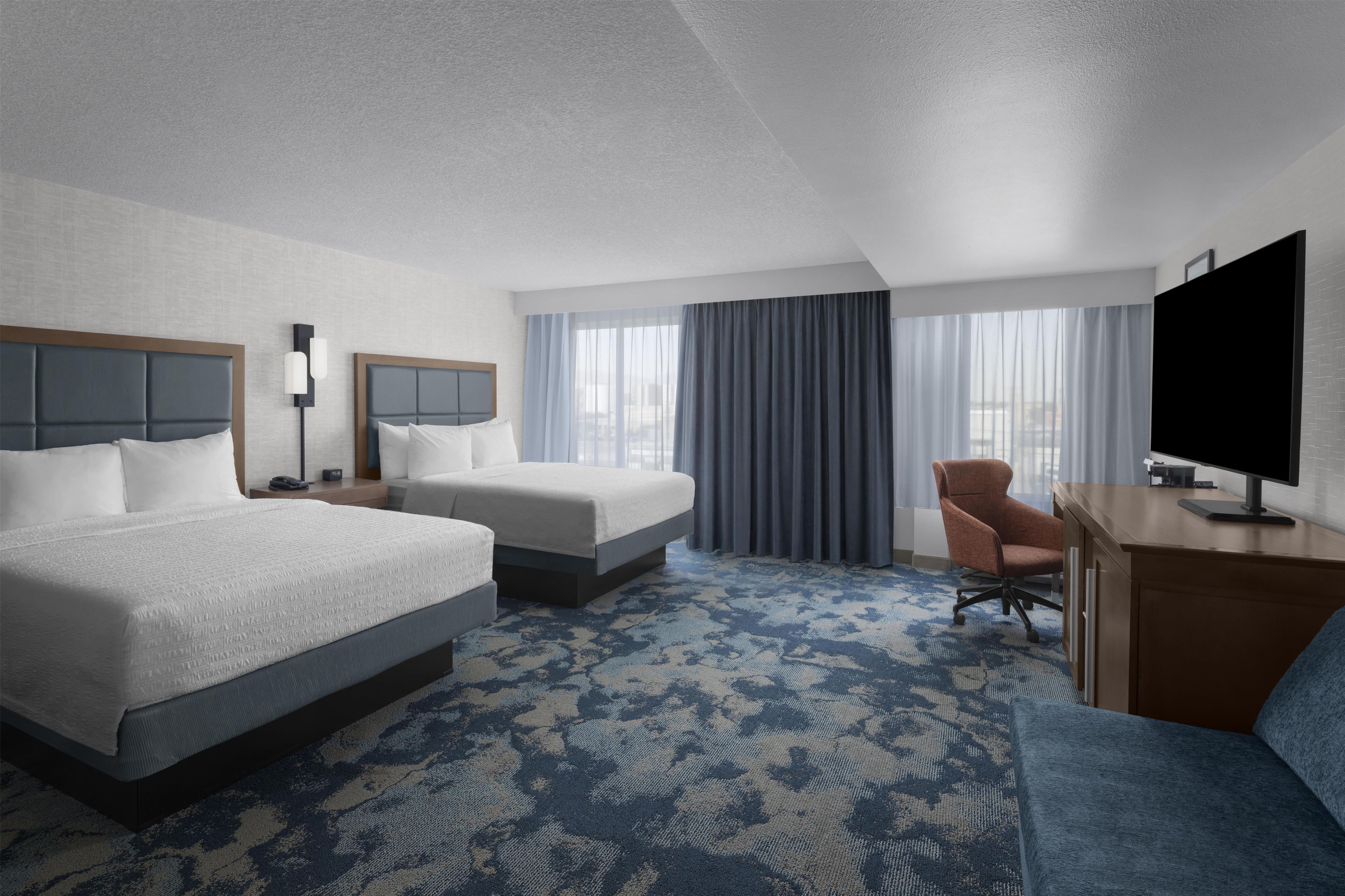 A hotel room with two beds, a desk, a chair, a TV, and a large window with curtains.