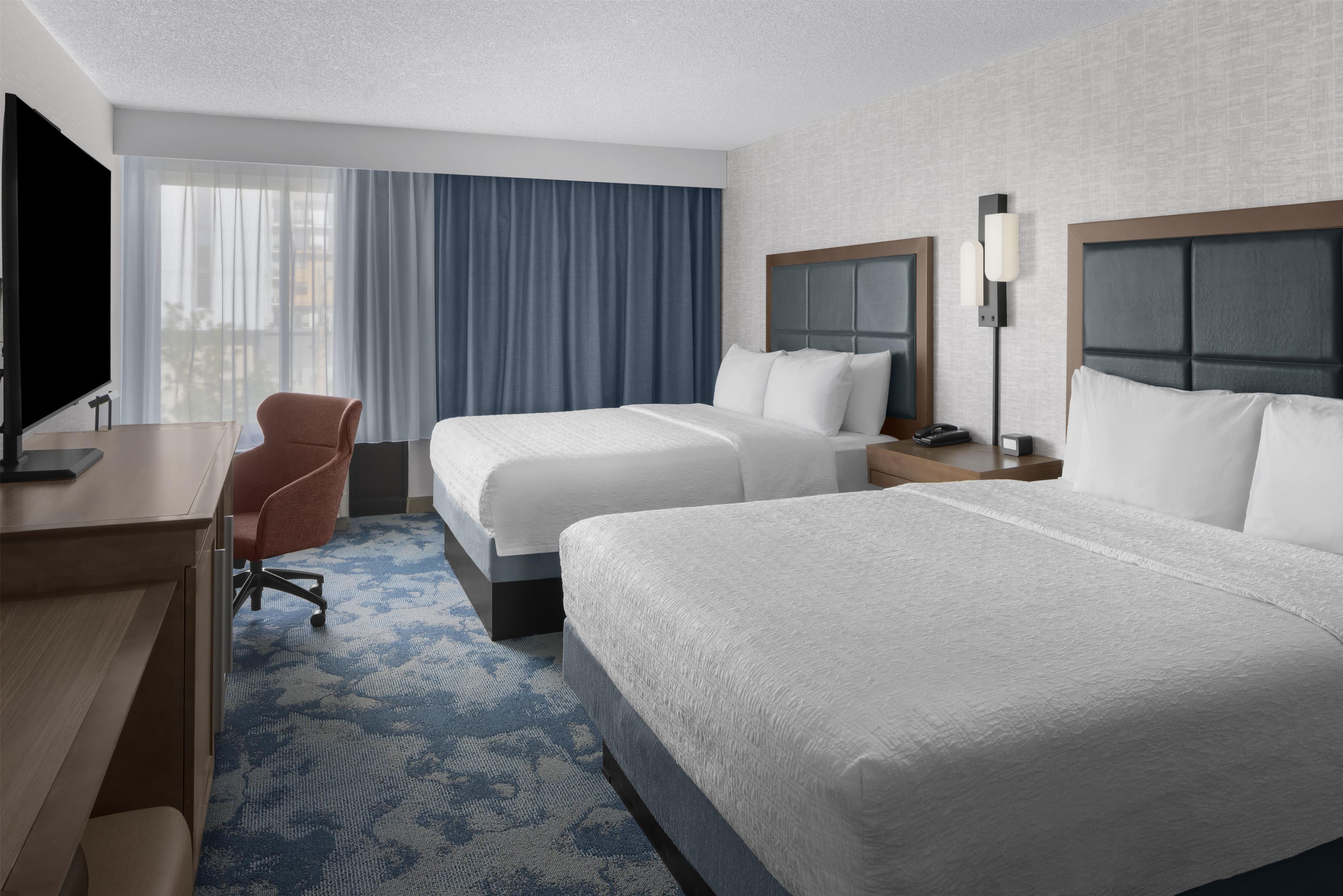 A hotel room with two beds, a desk, a chair, a TV, and a window with curtains.