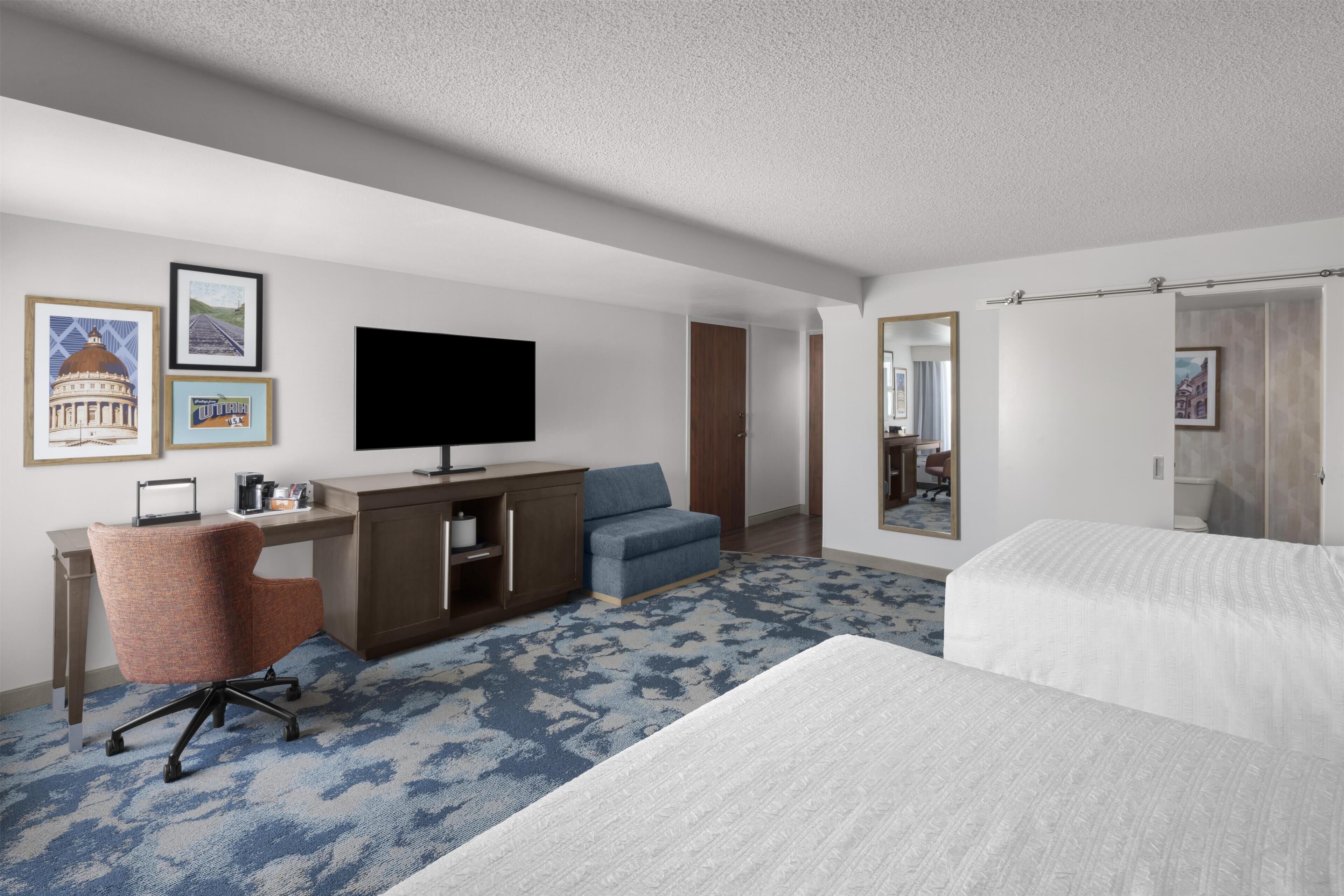 A hotel room with a bed, a desk with a chair, a television, and a large mirror.