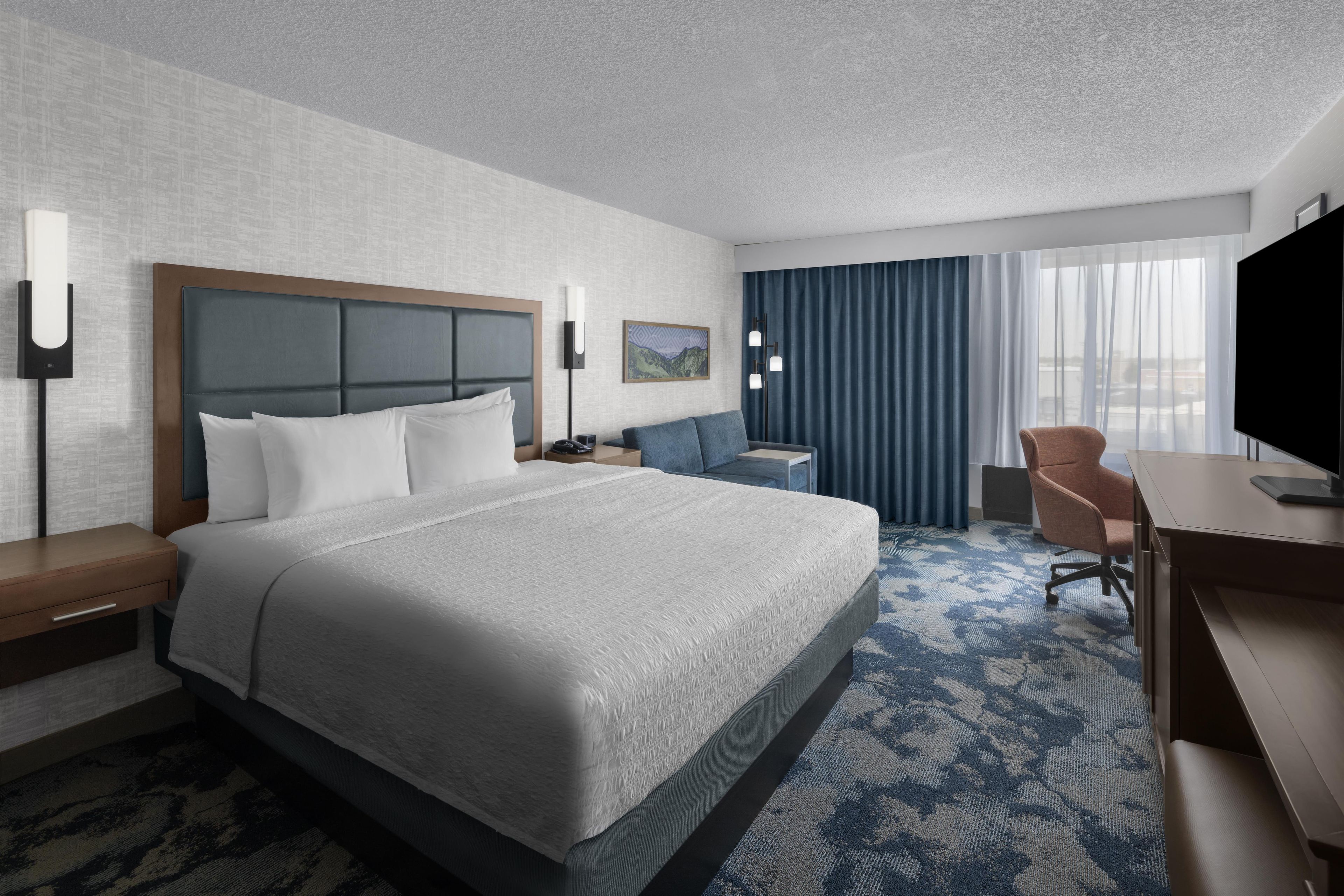 Habitación de hotel con una cama grande, un escritorio, una silla, un televisor y una ventana con cortinas.
