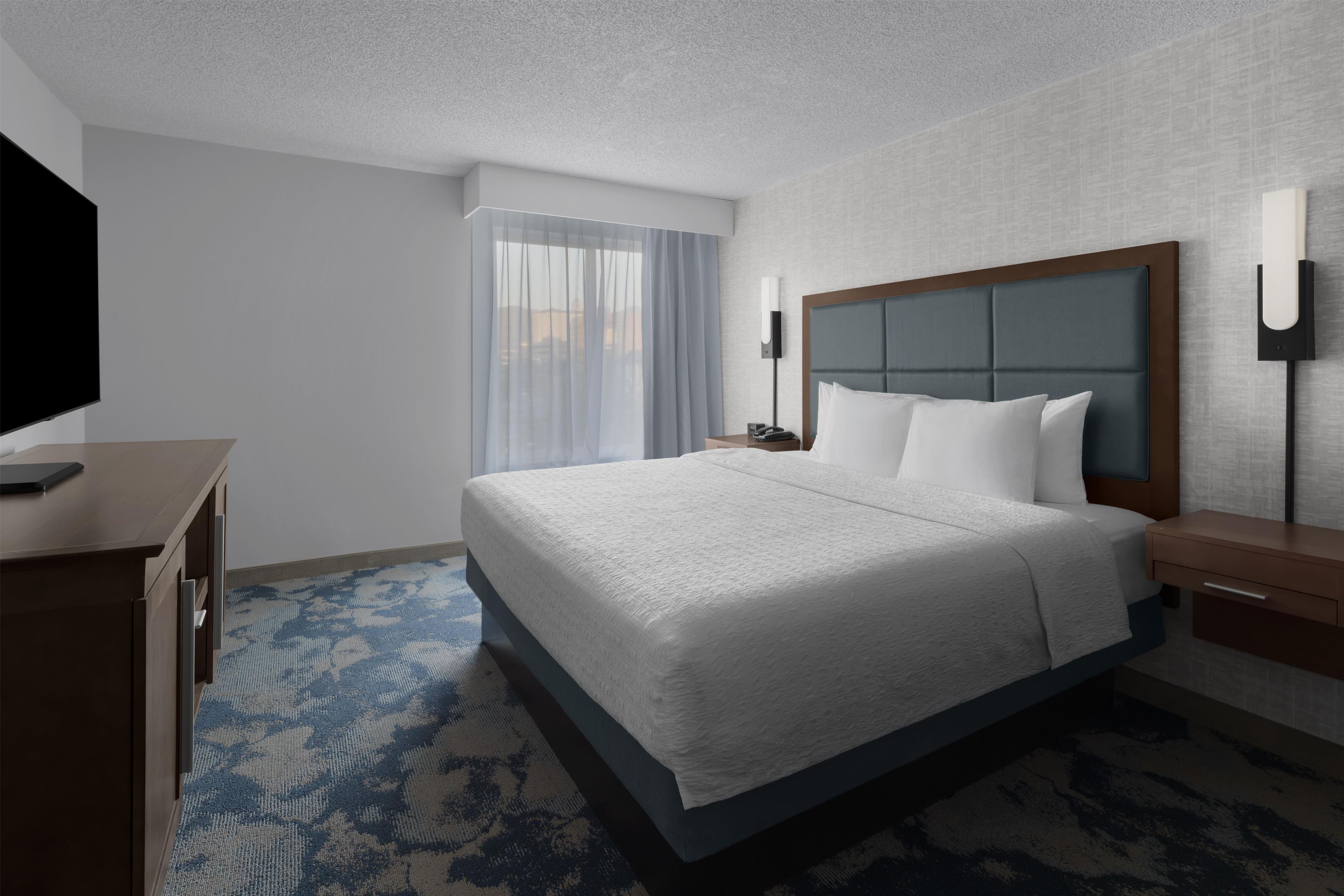 A hotel room with a bed, a desk, a TV, a window with curtains, and a wall-mounted light.